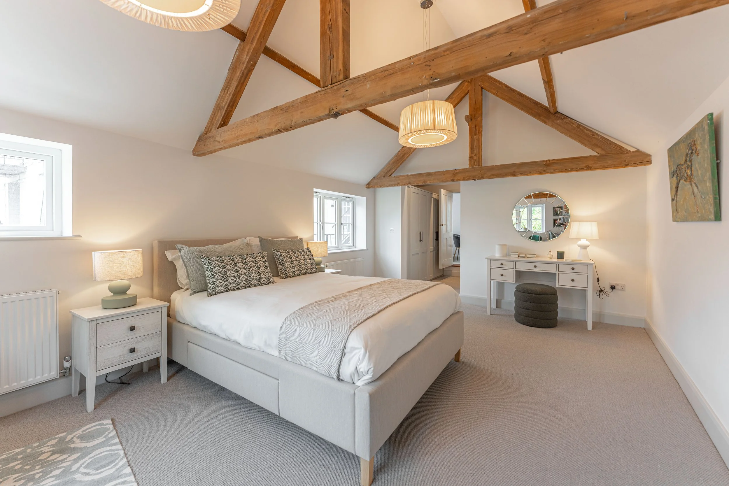 A cozy bedroom with a vaulted ceiling featuring exposed wooden beams. The room has a large bed with white bedding and decorative pillows, along with two small nightstands with lamps. There is a white dresser with a mirror, a small stool, and artwork 