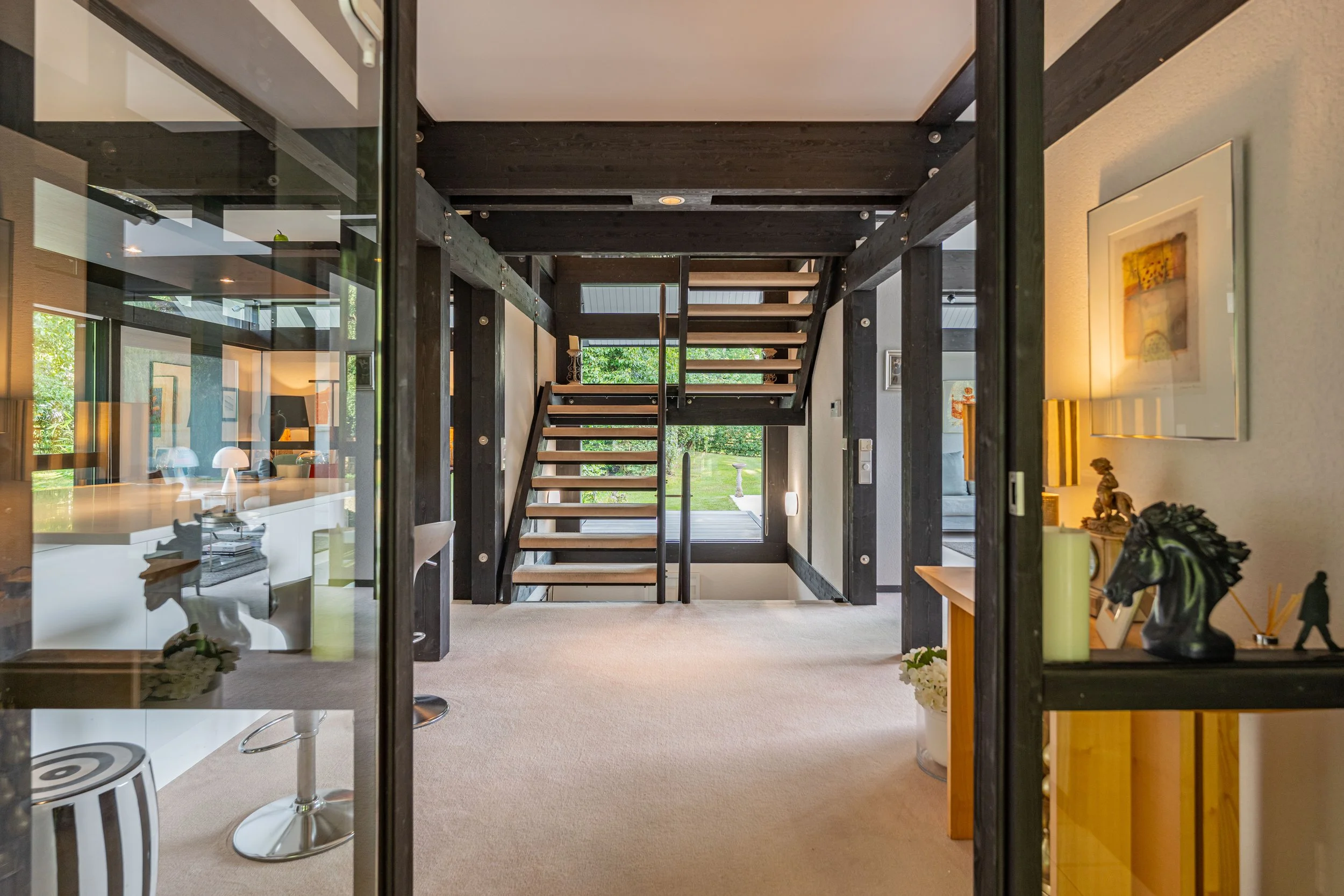 Interior view of a modern home taken from outside through glass doors, showing a staircase with dark wood and metal railings, and a living space with artwork, decorative items, and a dog sculpture.