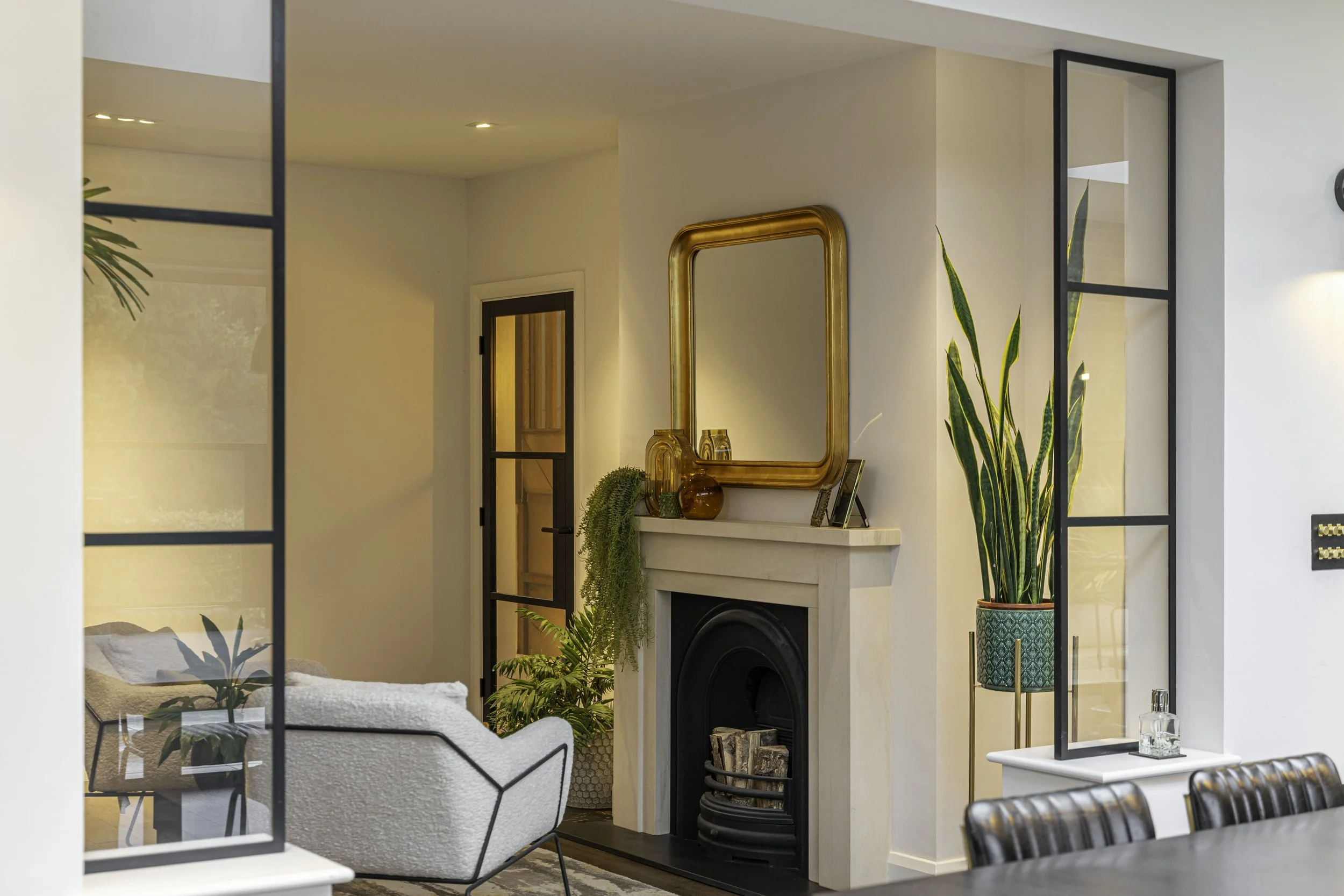 Living room with a white fireplace, a large mirror above it, and various decorative vases and plants. A modern armchair is partially visible on the left, and part of a dining table is seen at the bottom right.