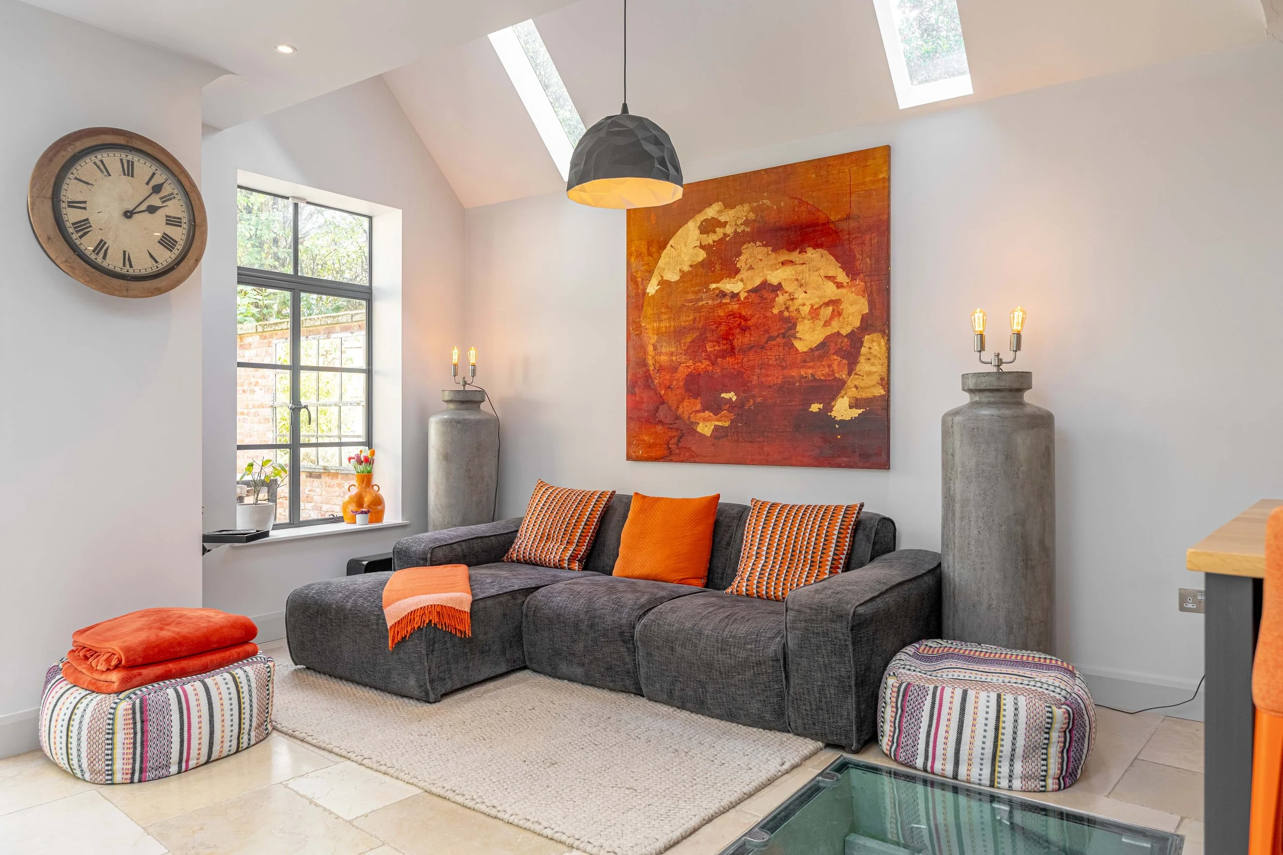 Living room with dark gray sectional sofa, orange pillows, orange and striped blanket, colorful pouf, abstract world map painting with orange and gold tones, concrete vases with lamps, skylights, window with a view of a brick wall and greenery, wall 