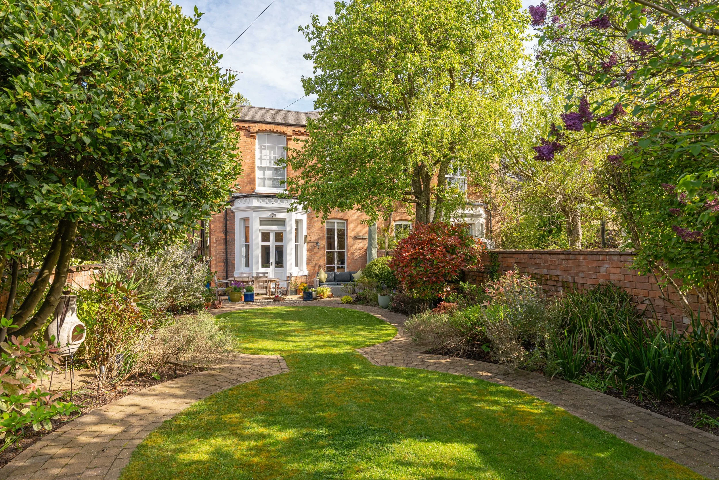 A lush garden with a curved brick pathway leading to a brick house with large windows, surrounded by trees and flowering shrubs.