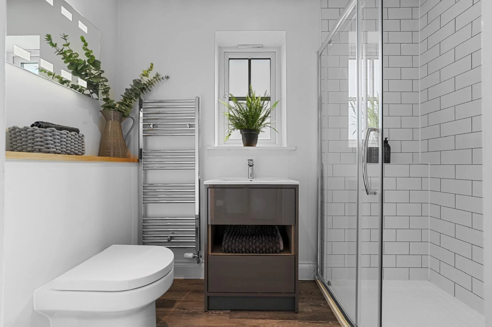 Modern bathroom with white walls, a small vanity with a sink beneath a window with a potted fern, a glass shower enclosure with white subway tiles, a chrome towel warmer, and decorative items like a large pitcher with greenery and neatly folded towels.