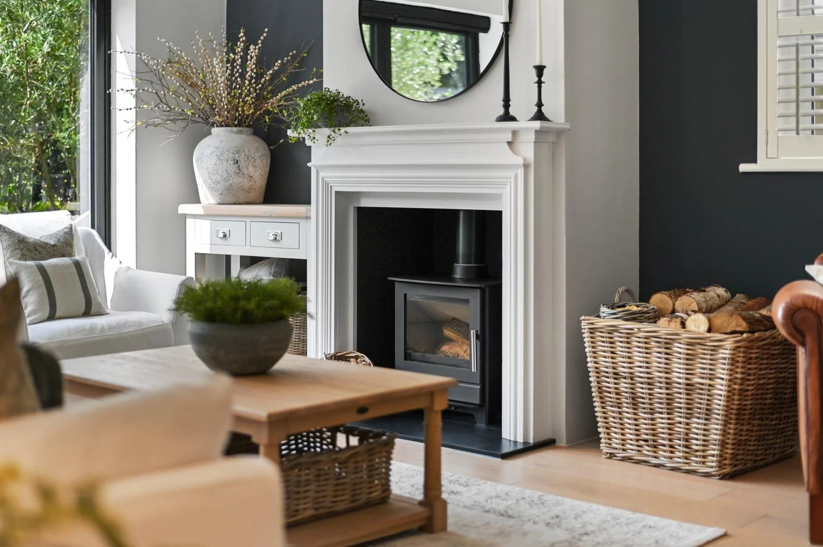 Living room with a white fireplace, black stove insert, and decorative mantel. There is a large vase with branches and a plant on the mantel. To the right, a basket filled with firewood. To the left, part of a white sofa with cushions, and a black framed window.
