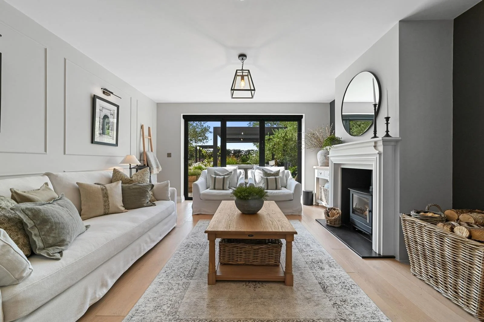 Bright living room with white sofa, armchair, wooden coffee table, fireplace, and large window showing outdoor greenery.