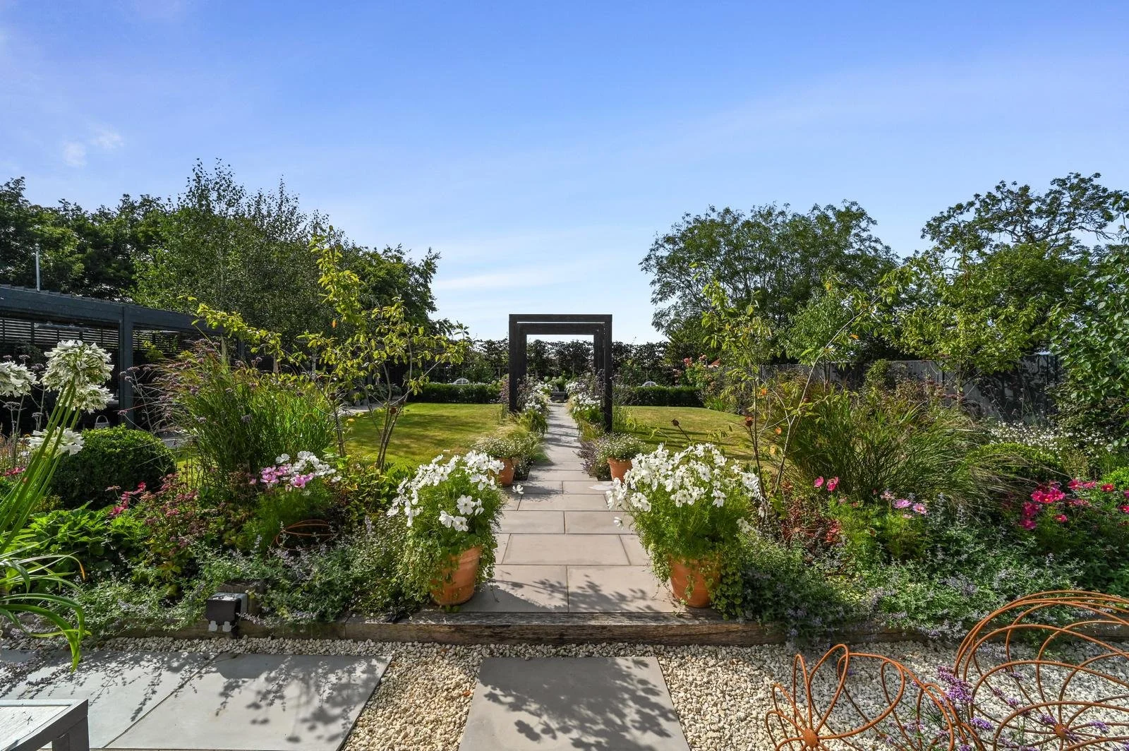A garden with a paved pathway leading through a black square archway, lined with potted white flowers, surrounded by green shrubs and trees under a clear blue sky.