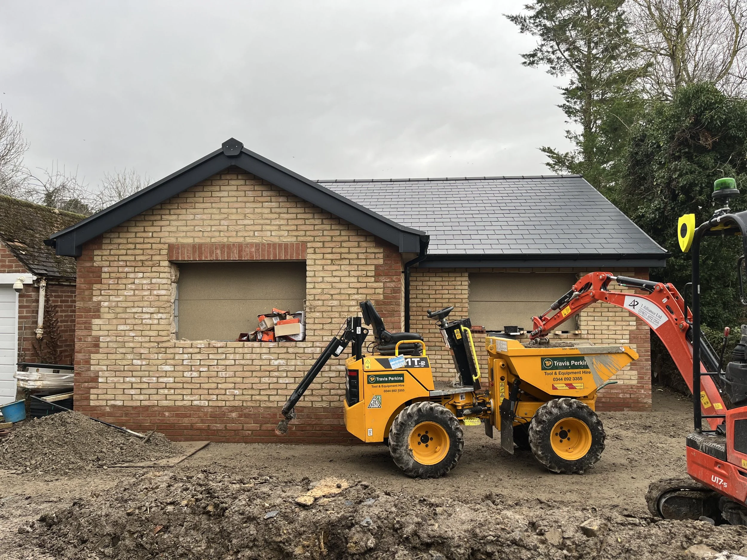 Construction site with a yellow mini loader and a small red excavator positioned in front of a brick building under construction, with construction materials inside the window opening.