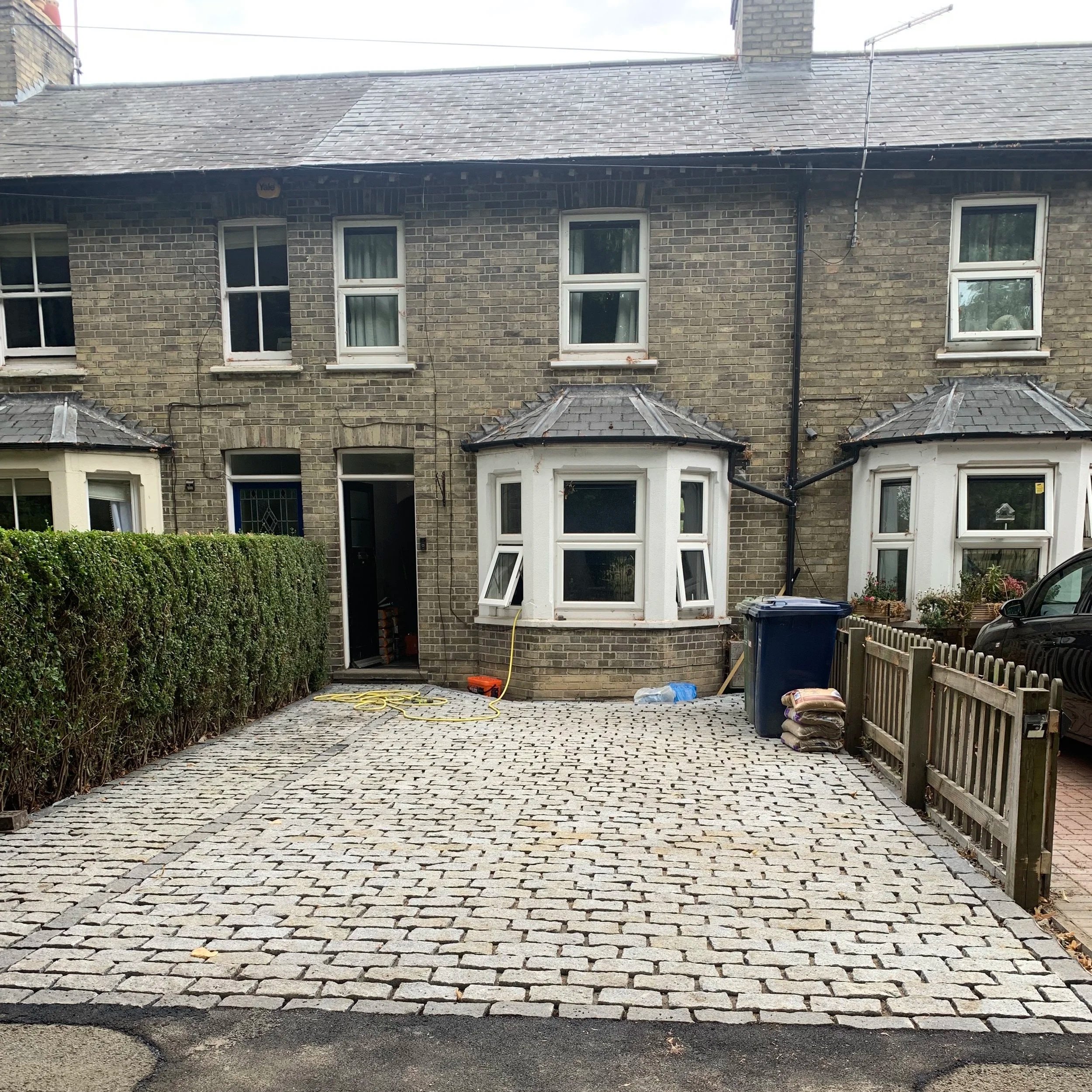 Front view of a brick row house with a paved driveway, and a small garden area with potted plants and a hedge on the left side.