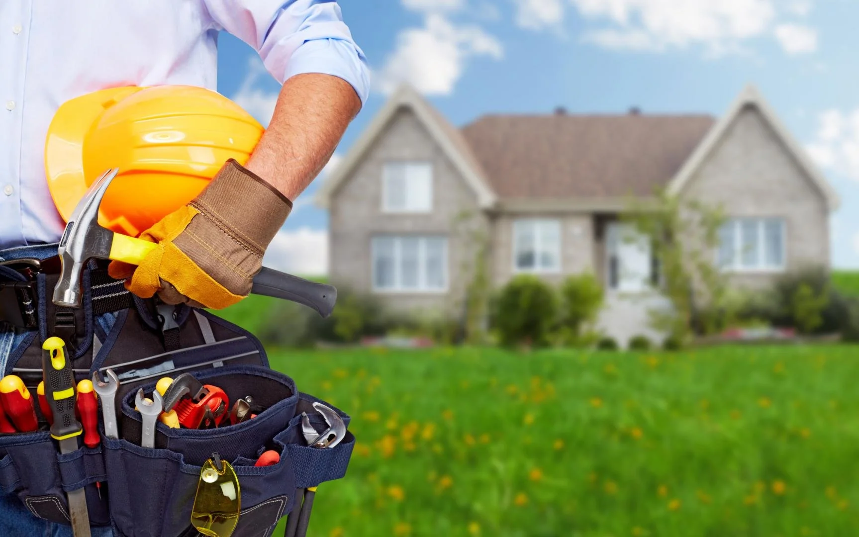 Close-up of a handyman with tools and a yellow safety helmet in front of a house on a green lawn.