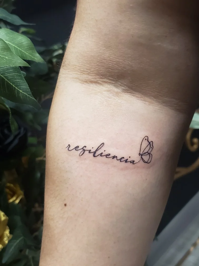 Text tattoo of the word Resiliencia with a butterfly at the end of the word.