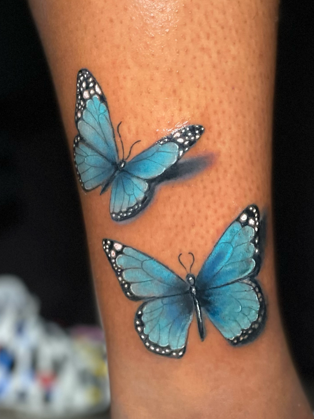 Vibrant tattoo of 2 blue butterflies with shadows below them.