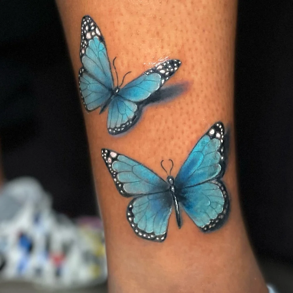 Vibrant tattoo of 2 blue butterflies with shadows below them.