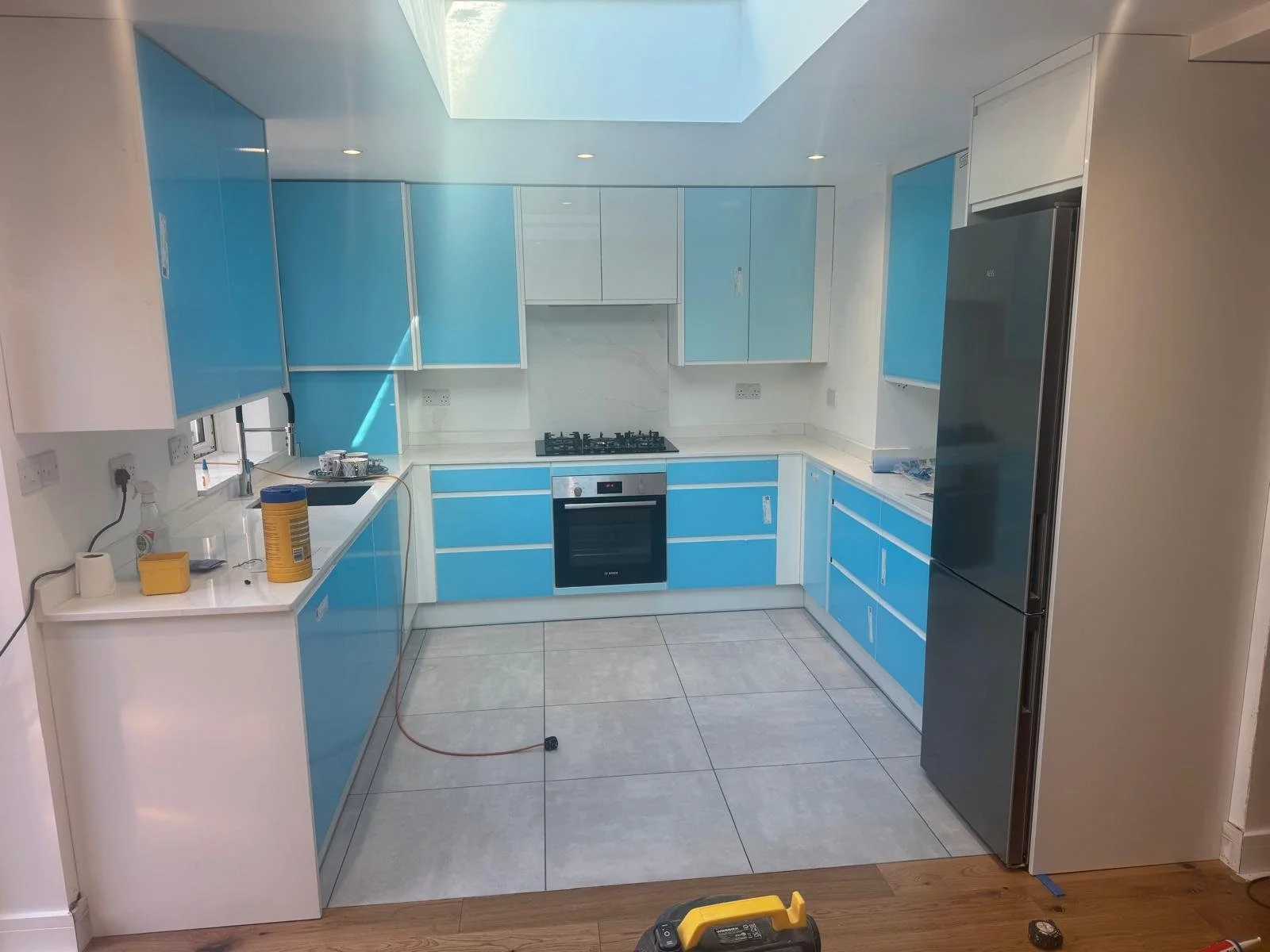 Modern kitchen with blue and white cabinets, stainless steel refrigerator, built-in oven, and gas stove. The kitchen is under renovation with tools and materials on the counter.