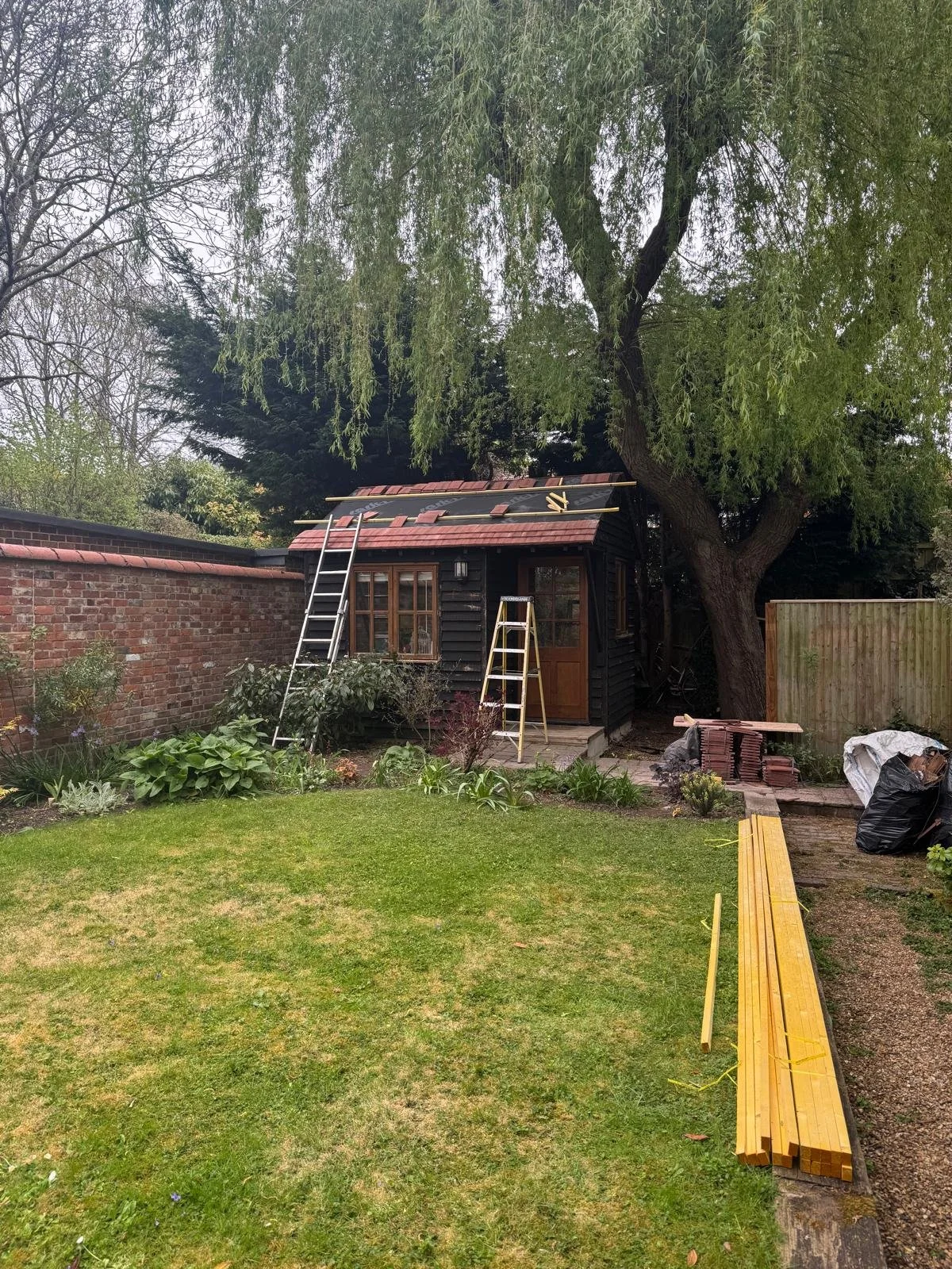 Backyard garden with a small black wooden shed under renovation, ladders leaning against it, surrounded by trees, plants, and a garden bed, with construction materials like stacked wooden planks and bricks nearby.