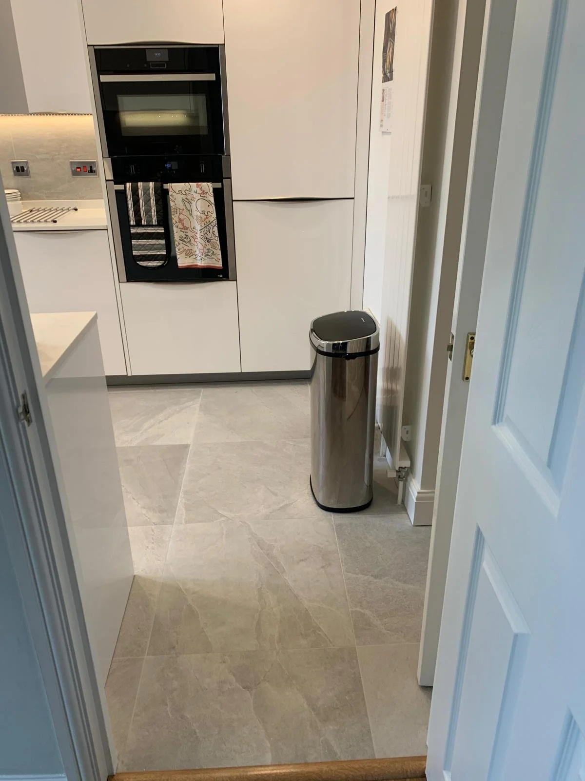 A modern kitchen with white cabinets, a built-in oven, a colorful towel hanging, a stainless steel trash can, and light-colored tiled flooring.