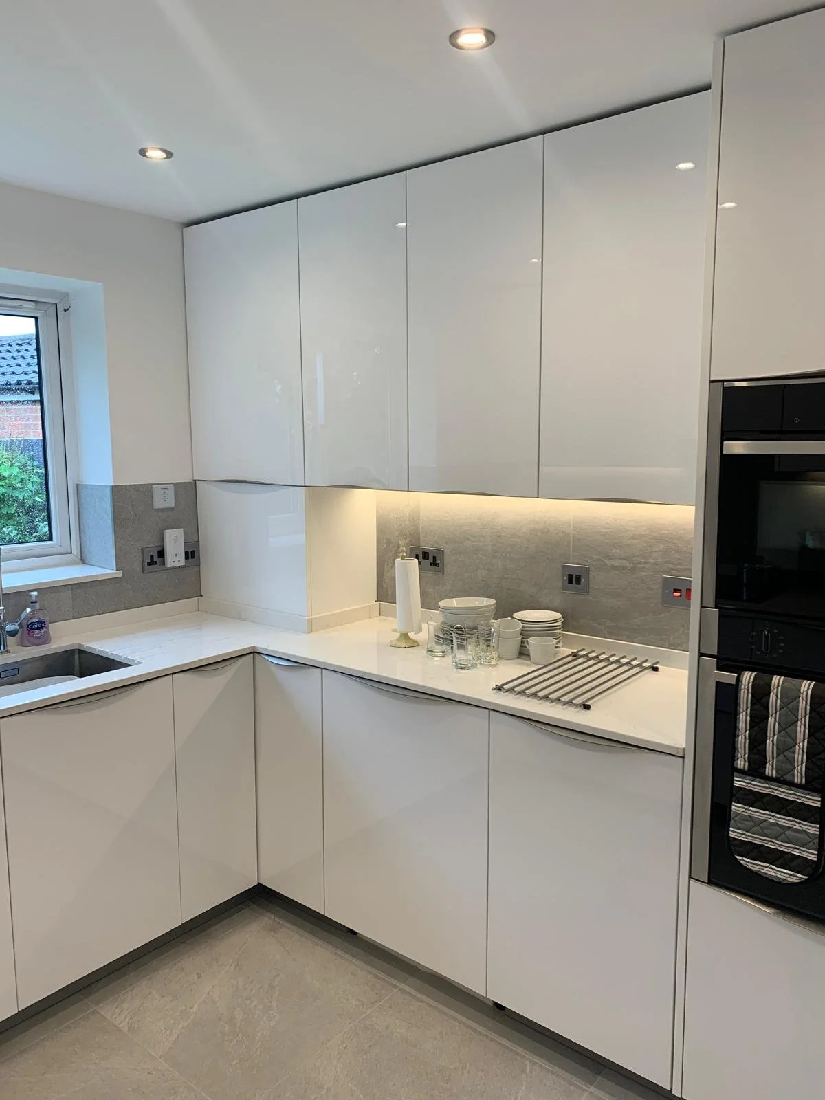 Modern kitchen with white cabinets, gray tiled backsplash, and a window. Kitchen counter has dishes, glasses, and a paper towel roll.