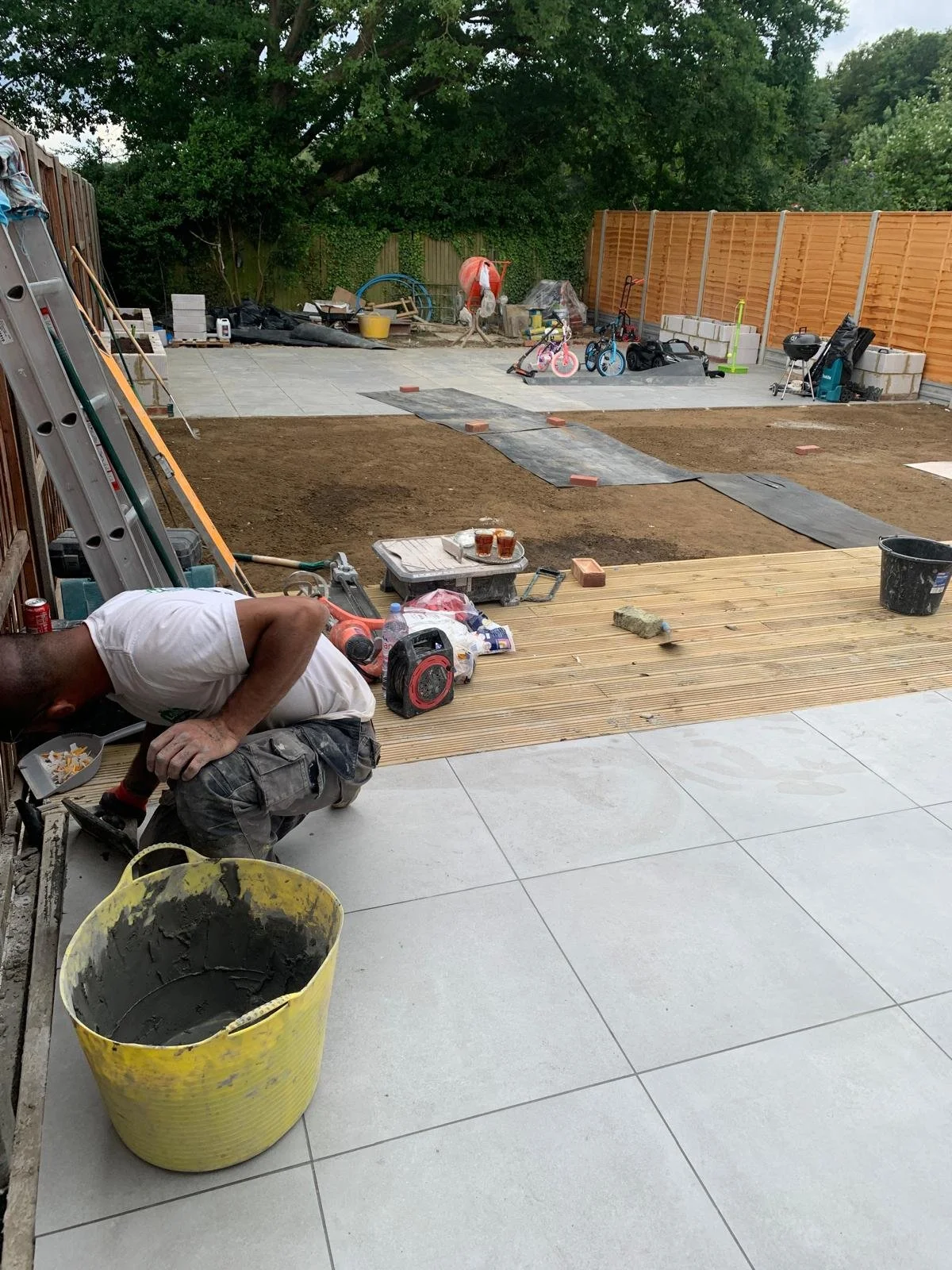 Backyard patio construction in progress with a worker laying large white tiles, construction tools, and materials scattered around. A partially completed patio area with exposed soil, children's bikes, and a wooden fence are visible in the background