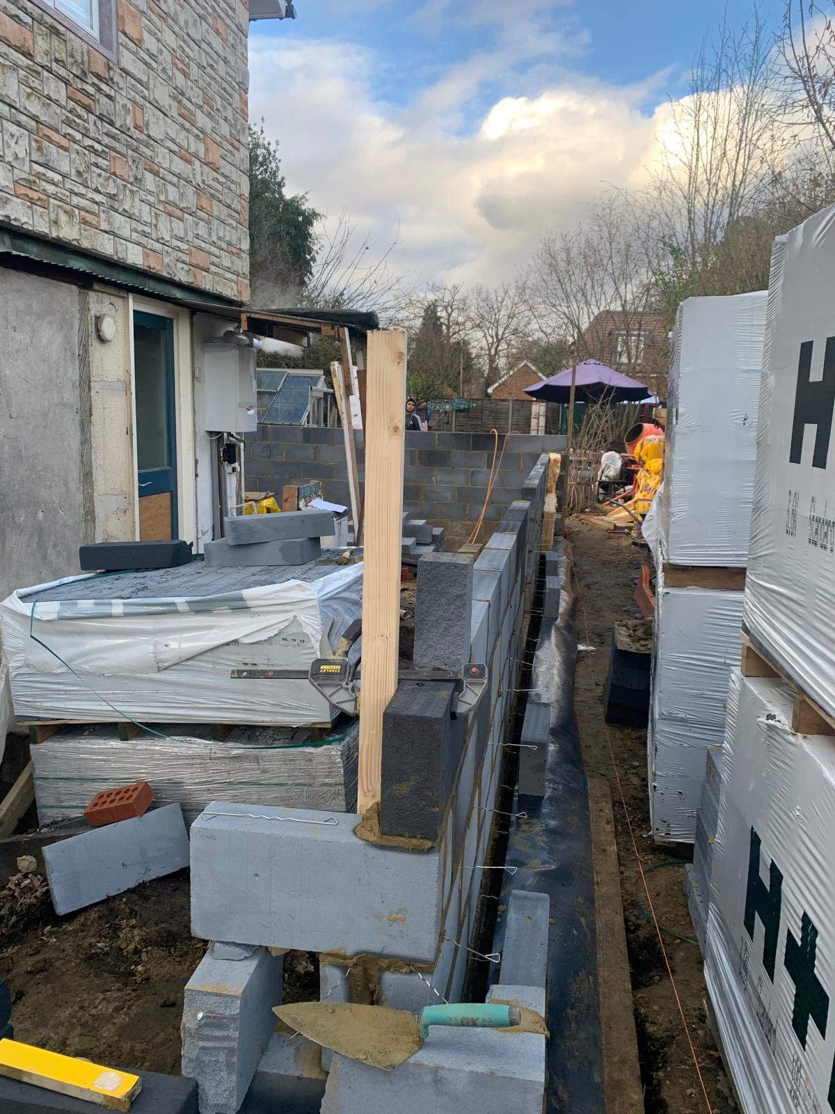 Construction site with a partially built stone wall, wooden posts, and stacks of insulation boards, with tools and construction materials nearby, in an outdoor residential area under a cloudy sky.