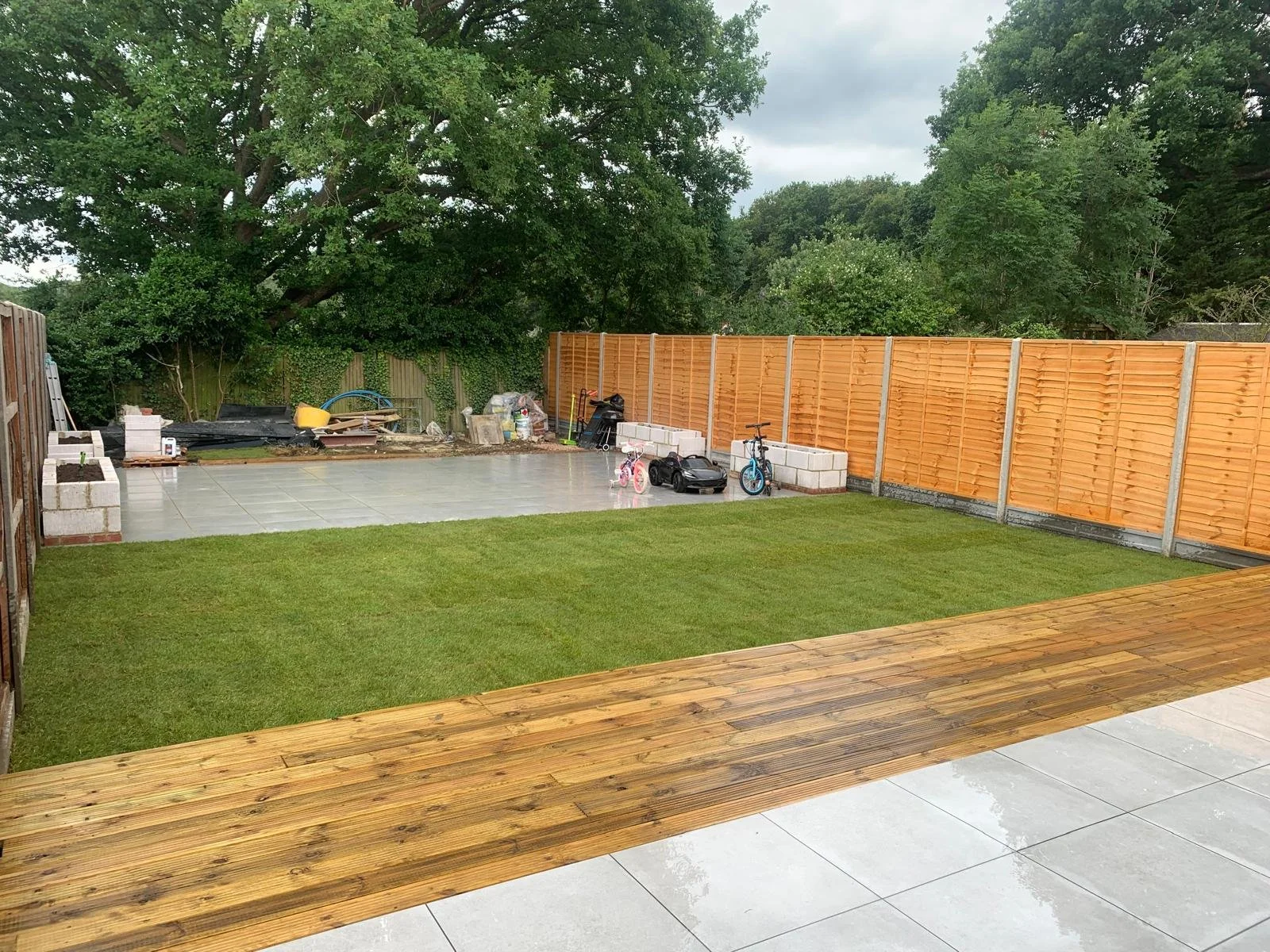 View of a backyard with a lawn, wooden deck, and a fenced area with construction materials and children's toys.