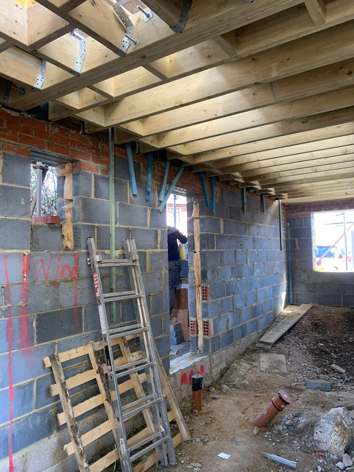Interior view of a construction site showing unfinished brick and concrete walls, wooden ceiling framing, scaffolding, and construction tools and materials on the ground.