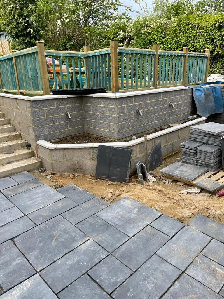 Under construction backyard with stone patio, brick wall, and wooden fence with stairs.