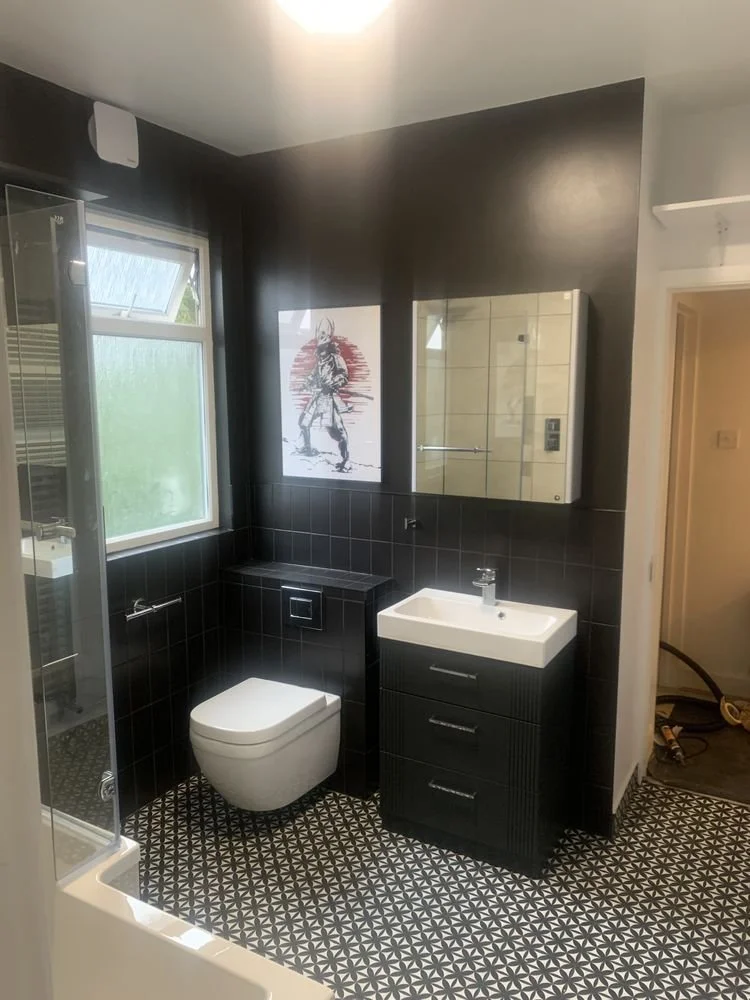 Small bathroom with black tile walls and patterned black and white floor. Contains a window, a wall-mounted toilet, a black vanity with a white sink, and a mirror with storage. Artwork of a Native American man hangs on the wall.