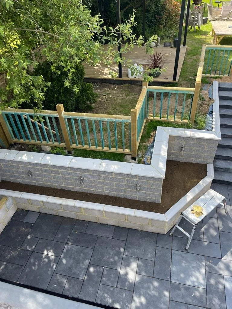 Construction of a brick and concrete retaining wall with a wooden fence on top, adjacent to a patio with black tile flooring, and a small silver stool with a yellow cloth on it; in a backyard with grassy and landscaped areas.