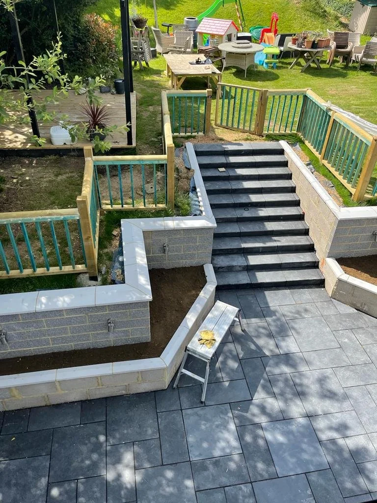 A backyard with a tiled patio, stairs, and newly built brick walls. There are garden fences with blue and green posts, a wooden outdoor table, various toys, and garden beds with plants and gardening tools.