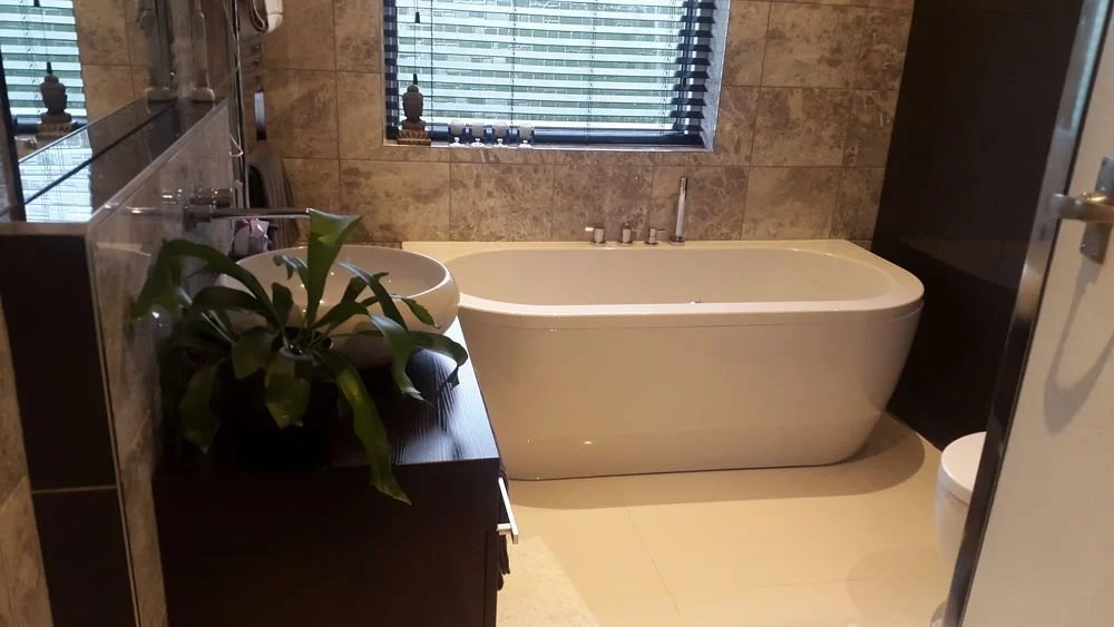 A modern bathroom with a bathtub, a plant on a dark wooden countertop, a sink, a mirror, and a window with blinds.