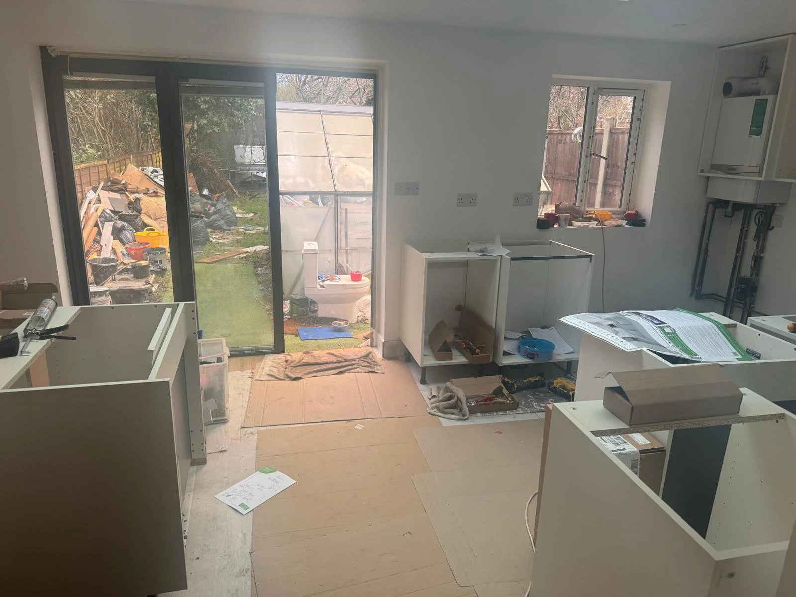 Interior view of a kitchen under renovation with partially assembled cabinets, tools, construction materials, and an open sliding door leading to a garden with debris pile outside.