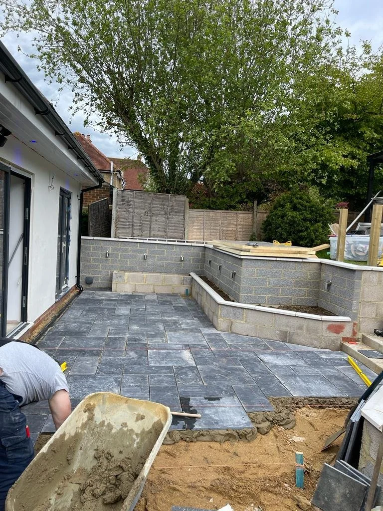 Construction site with a partially built brick wall, tiled patio, and a worker laying tiles in a backyard.