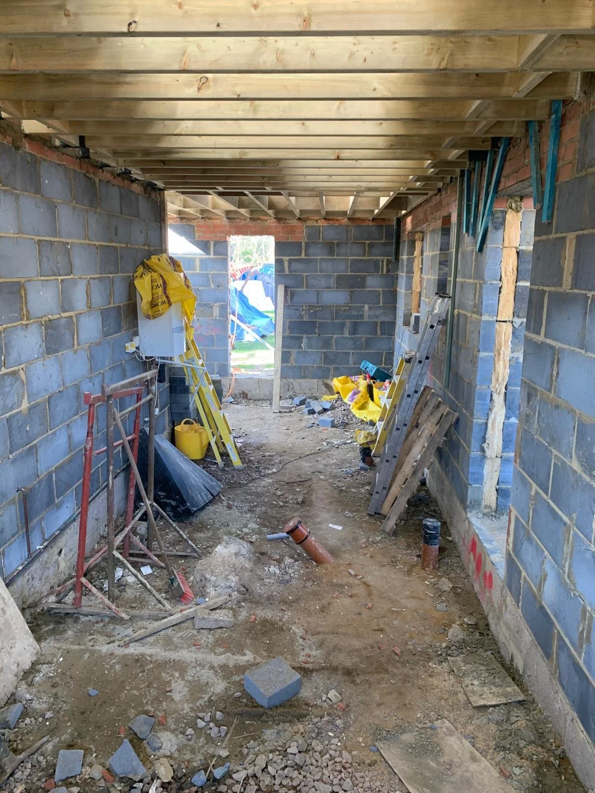Interior view of a house under construction with brick walls, a wooden ceiling structure, and construction tools and materials scattered on the dirt floor.