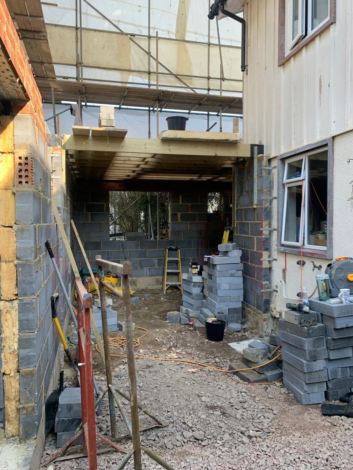 Construction site of an extension or new building with brick walls, scaffolding, construction tools, stacked bricks, and an unfinished interior space.