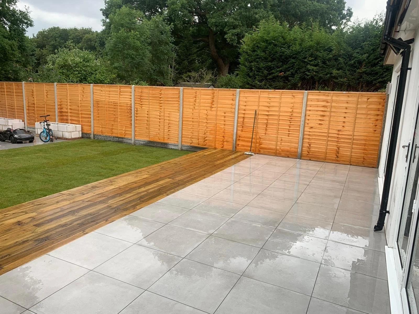 A backyard patio with large light gray tiles, a small section of wet wood decking, a green lawn, a wooden fence, a broom, a blue bicycle, black toy car, and trees in the background.