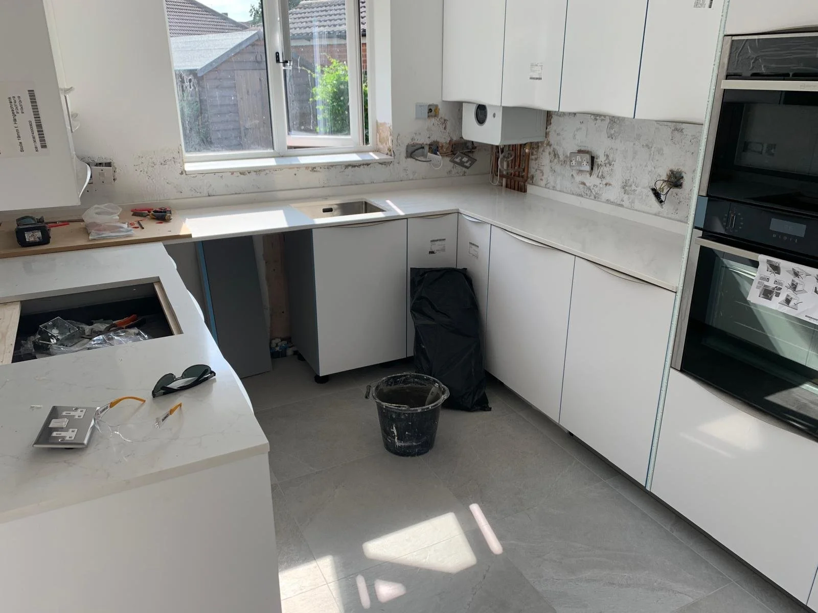 A kitchen under renovation with white cabinets, a window above the sink, and construction tools and materials scattered around.