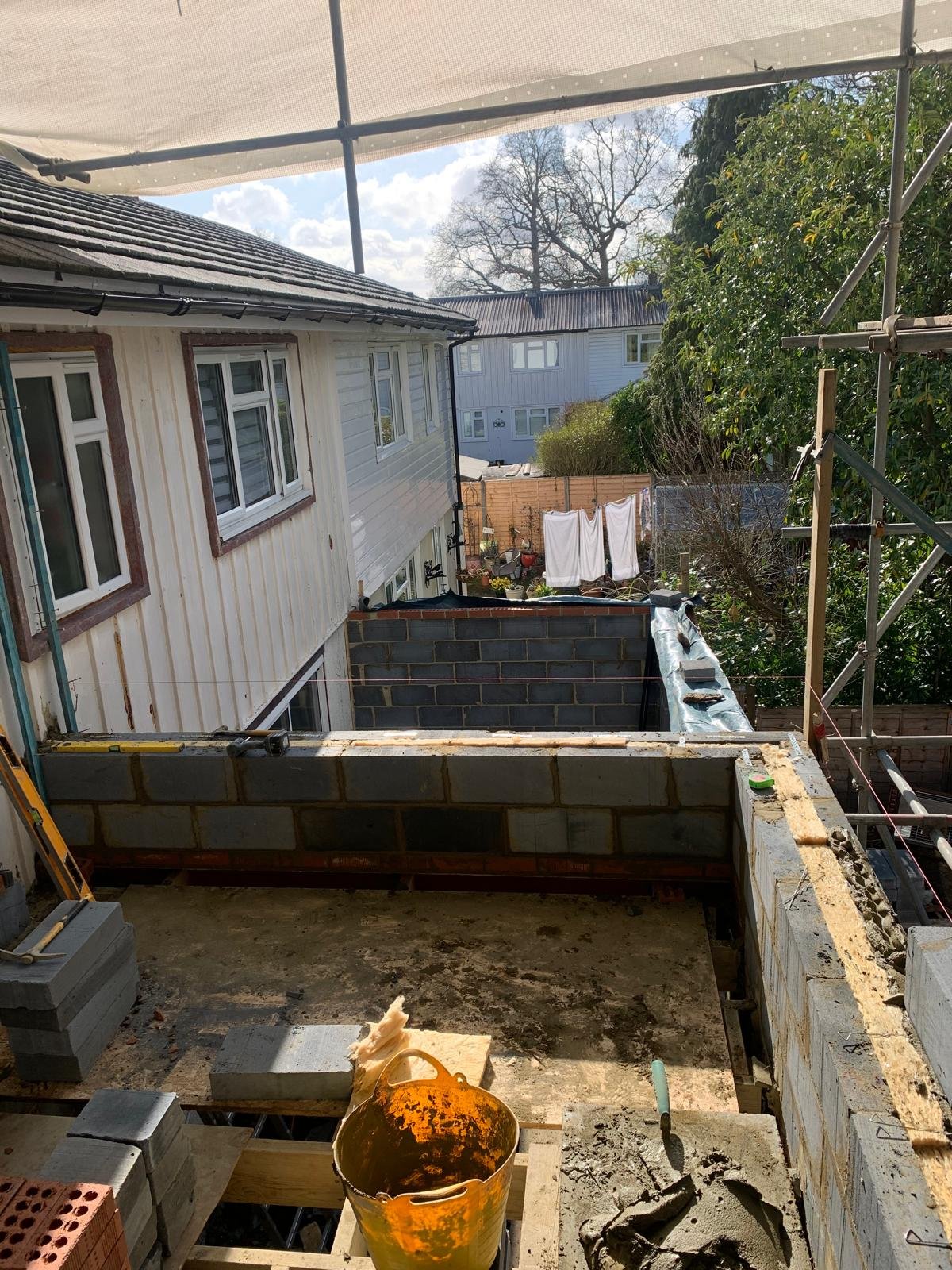View from a construction site on a residential building under renovation, with wall partially built, construction tools, and a bucket in foreground, neighboring houses and a sunny sky in background.