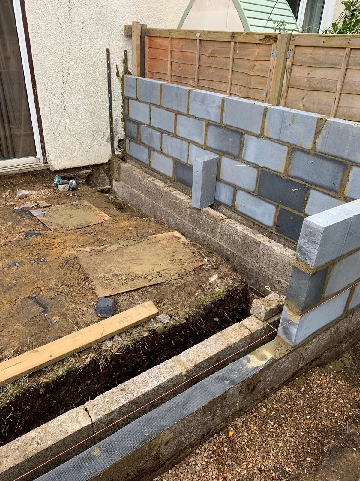 Construction site showing a partially built cinder block wall next to a house, with a wooden fence in the background, and construction tools in the corner.
