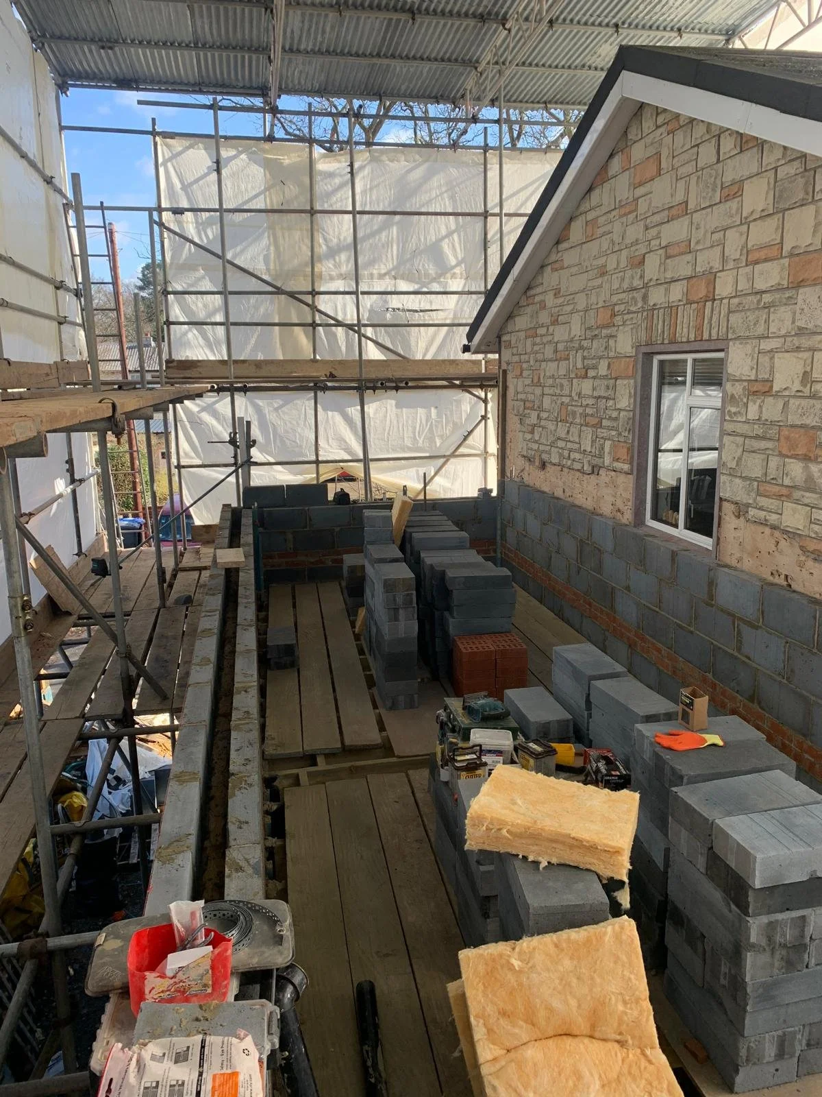 Construction site of a house with scaffolding, brick and stone walls, and wooden flooring, showing ongoing masonry and construction work.