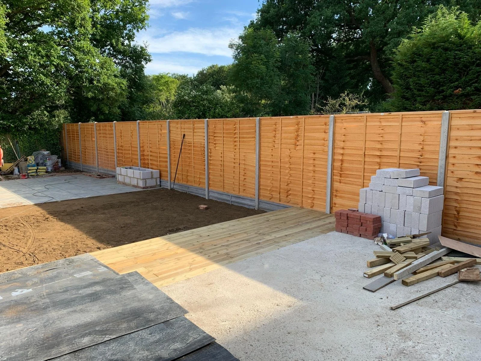 Backyard under construction with a new wooden fence, stacks of bricks, wooden planks, and a partially paved ground area.