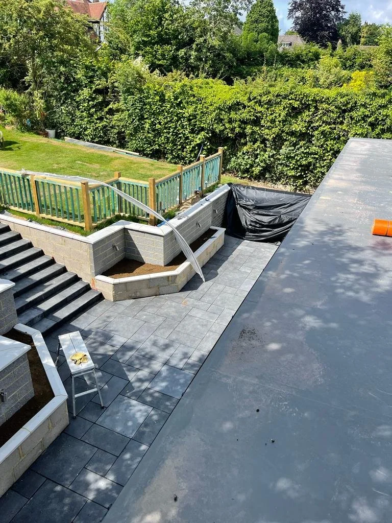 Construction of a backyard patio with brick retaining wall, water feature, and new paver flooring, with trees and a lawn in the background.
