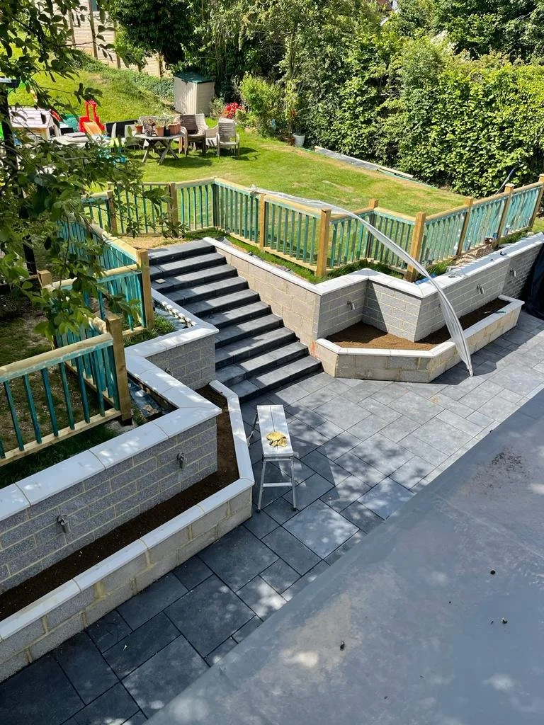 View of a backyard patio with newly constructed brick and cinder block retaining walls, a set of stone stairs, and wooden fencing. There are garden beds, a lush green lawn, a table with chairs and outdoor toys in the background.