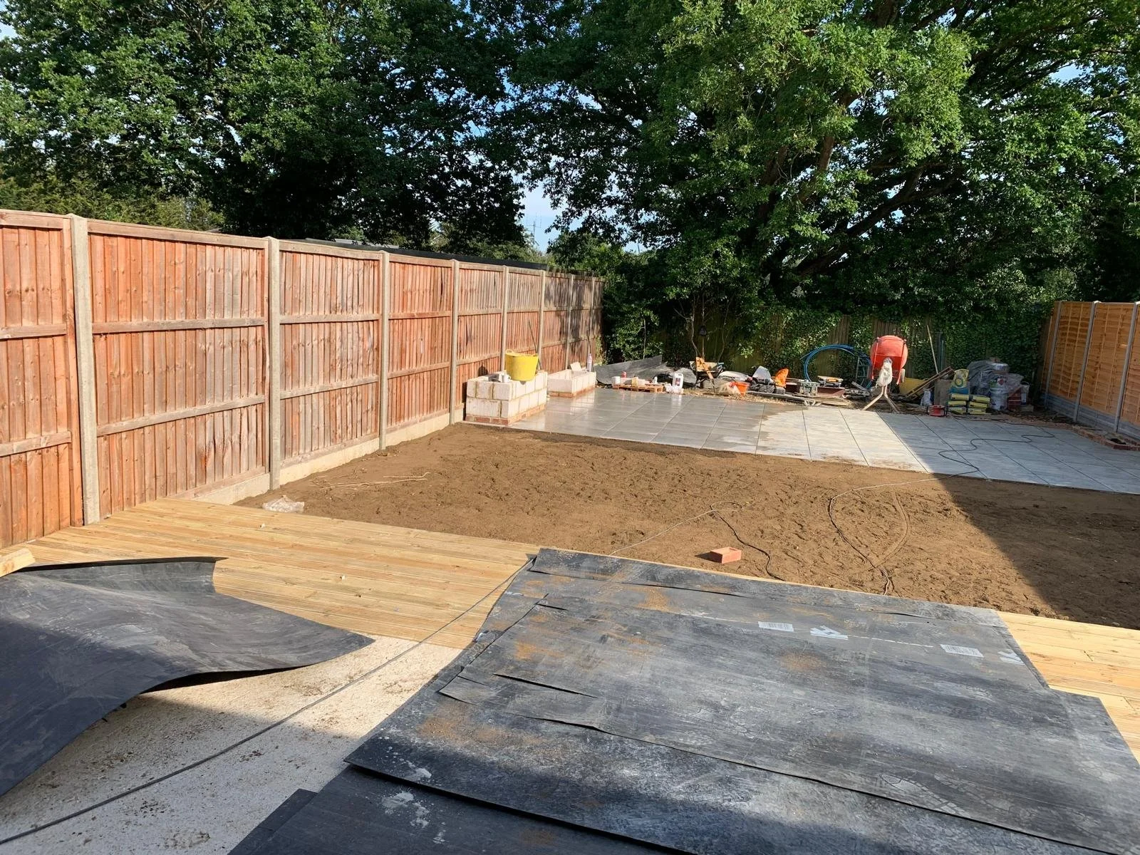 Backyard under construction with a newly installed wooden deck on the left, tiling on the right, and a partially leveled dirt area in the middle. Wooden fence surrounds the yard, and construction tools and materials are scattered around.