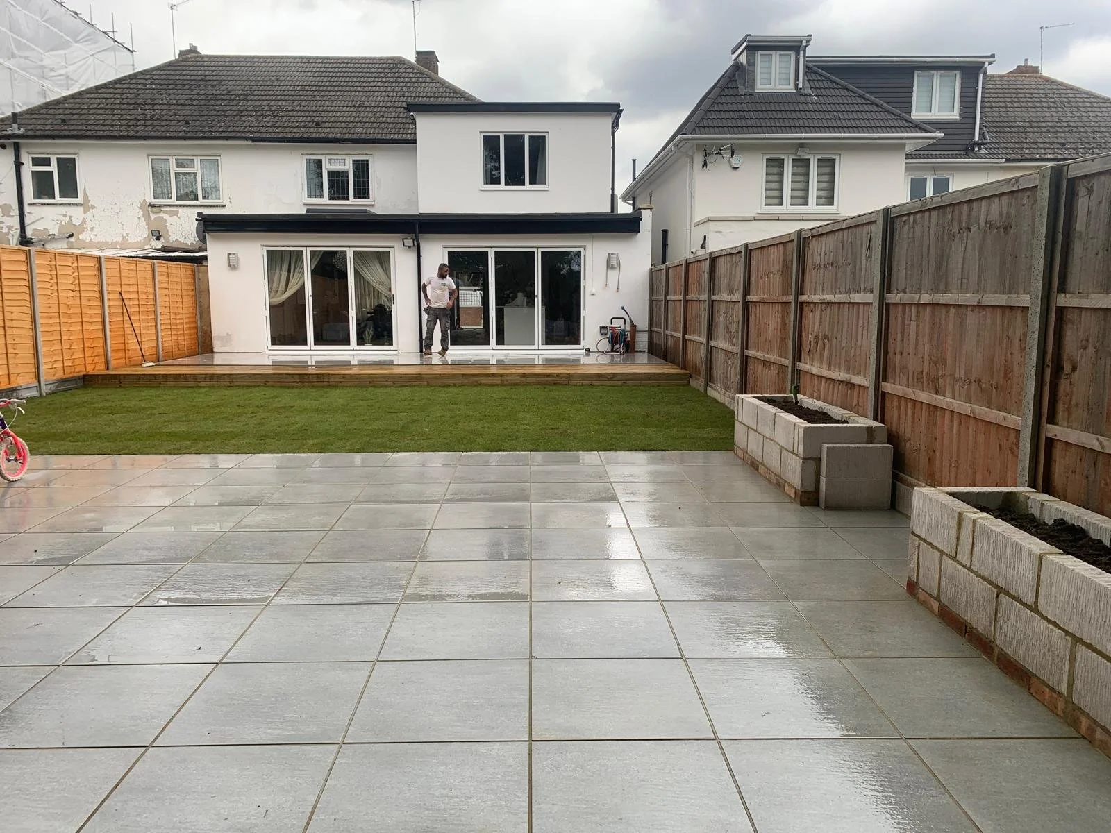 Backyard with a concrete patio, a small patch of grass, and a newly built deck area. Fences surround the yard, and there are houses in the background. A man is standing on the deck, and a pink bicycle is on the left.