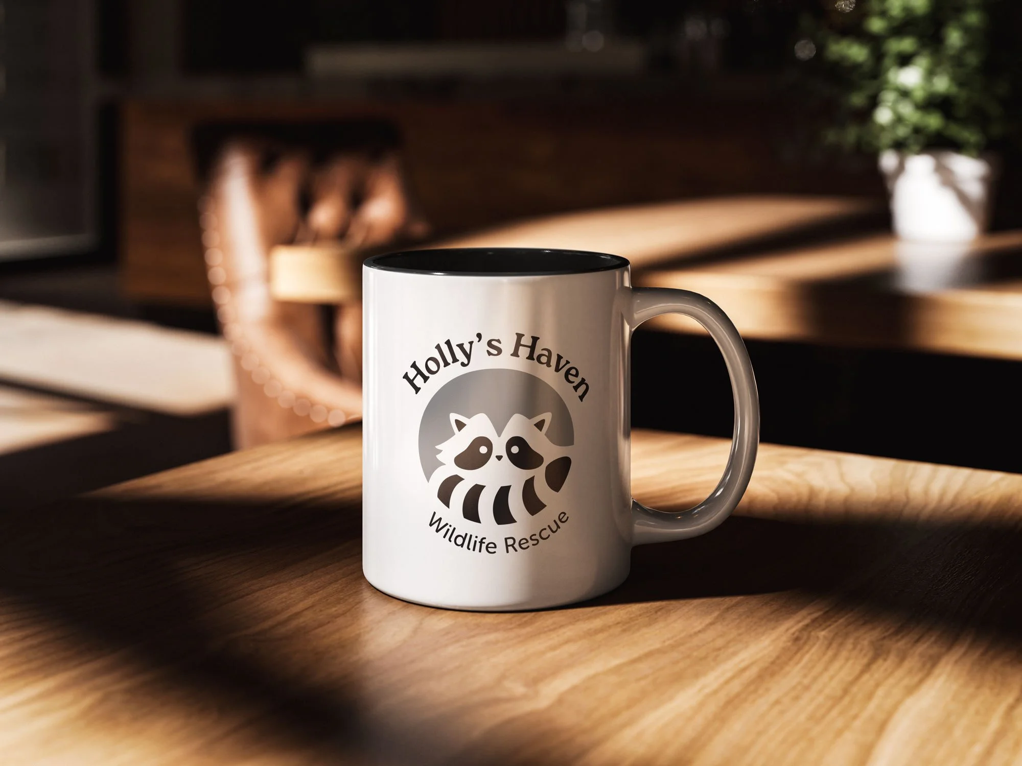 White ceramic coffee mug with black interior sitting on a wooden table in natural lighting. The mug features "Holly's Haven Wildlife Rescue" text and logo showing a stylized raccoon design in gray tones. Blurred background shows a dining room with wo