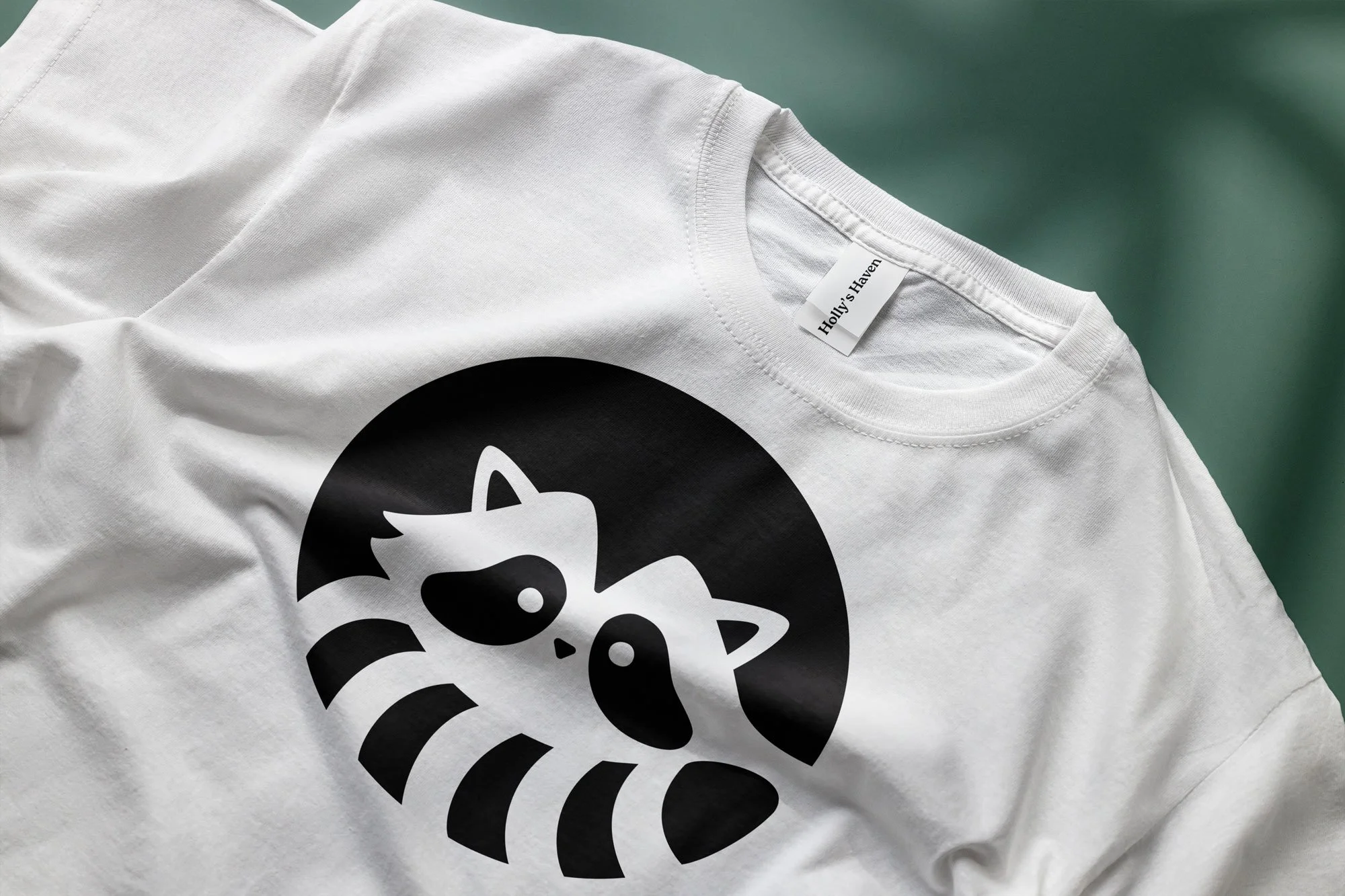 White t-shirt laid flat showing a black circular design of a stylized raccoon logo. A clothing tag visible at the neckline reads "Holly's Haven." The shirt is photographed against a muted teal-green background