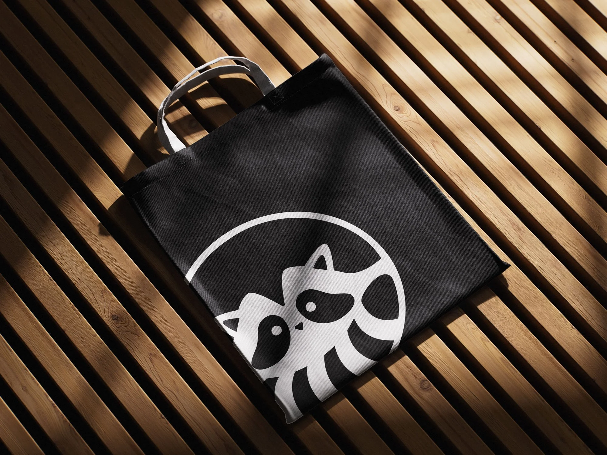 Black canvas tote bag lying on wooden slat surface featuring a white printed design of a stylized raccoon inside a circular frame, photographed from above in natural lighting.