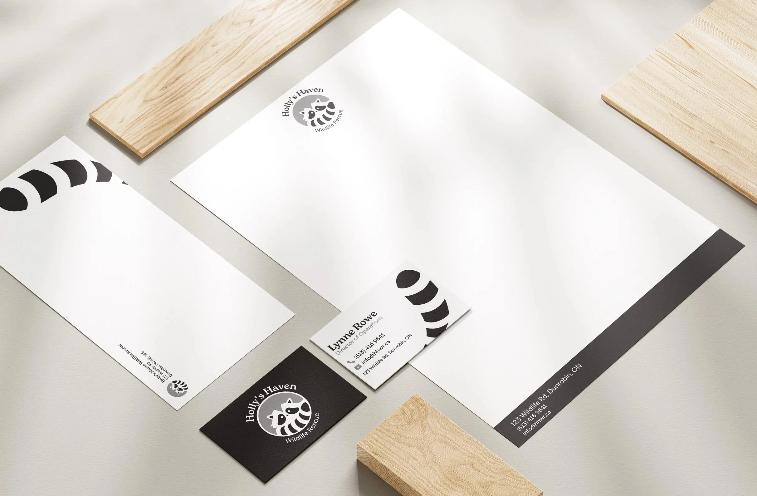 Flat lay of Holly's Haven Wildlife Rescue stationery mockup featuring letterhead, business cards in black and white, envelope, and decorative wood elements on a gray surface with wood planks.
