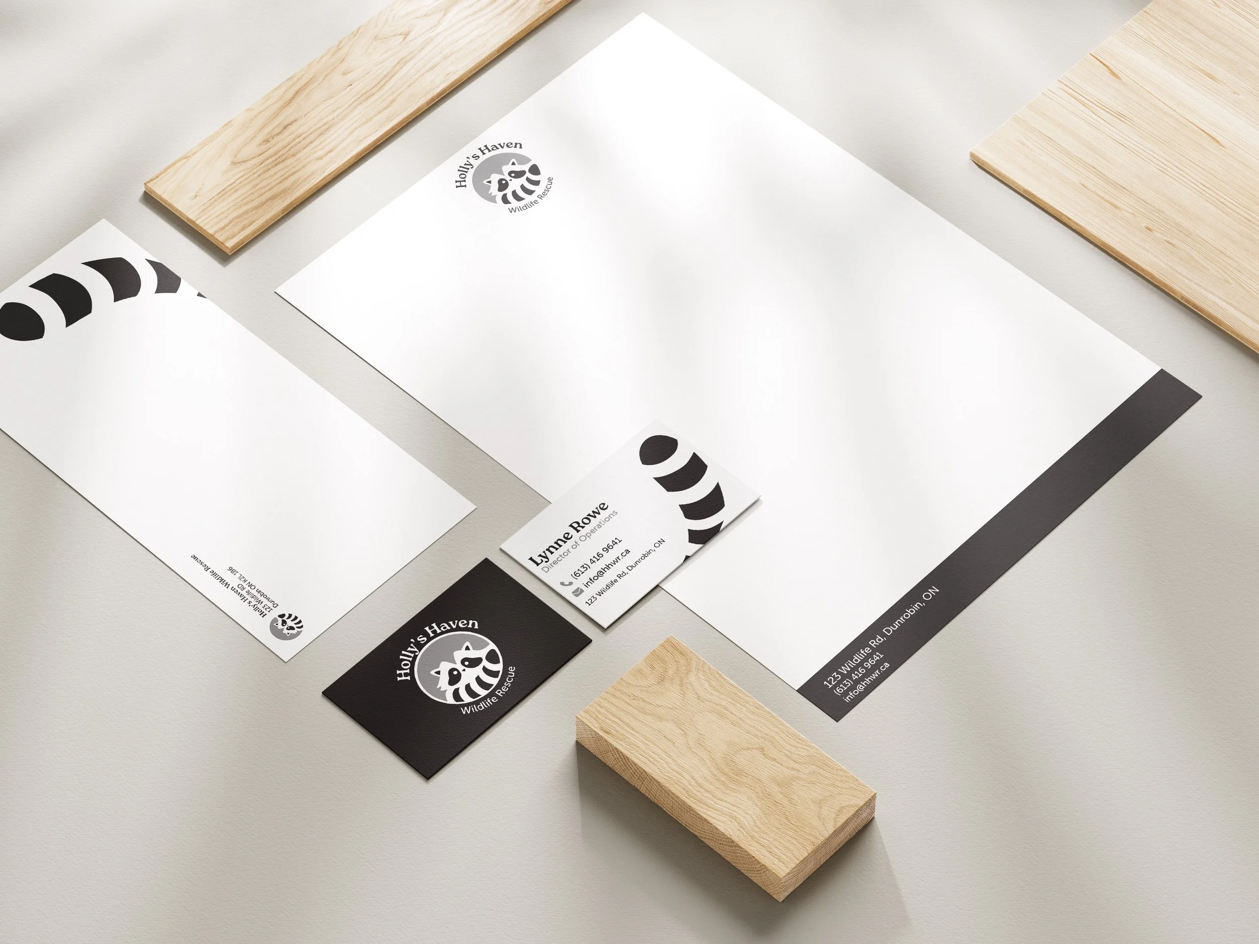 Flat lay photography of Holly's Haven Wildlife Rescue brand stationery mockup on a light gray surface with natural wood planks. The composition includes white letterhead with the raccoon logo at top, a white business card for Lynne Rowe showing conta