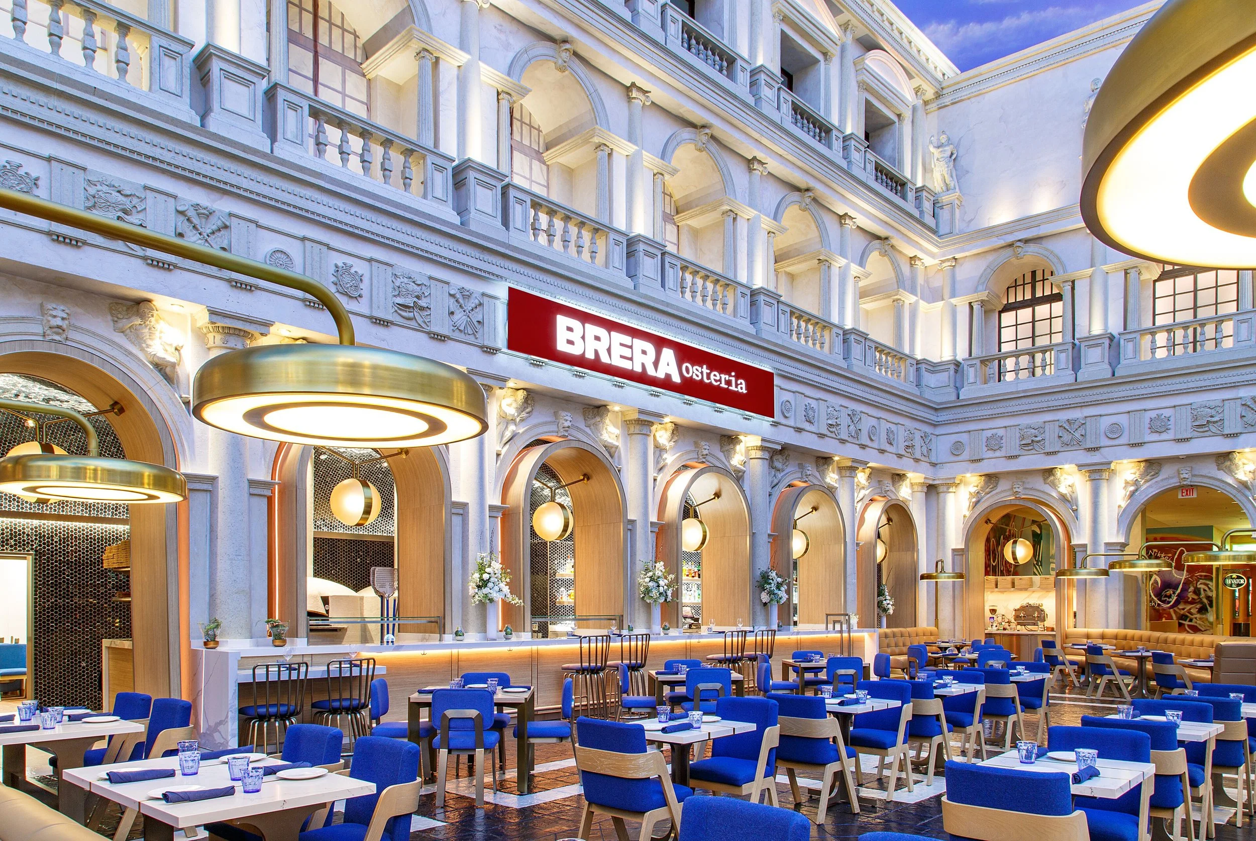 Elegant restaurant interior with ornate white architecture, arched windows, and gold lighting fixtures. Blue chairs surround white tables set for dining, with flowers and the sign 'BRERA Osteria' visible.