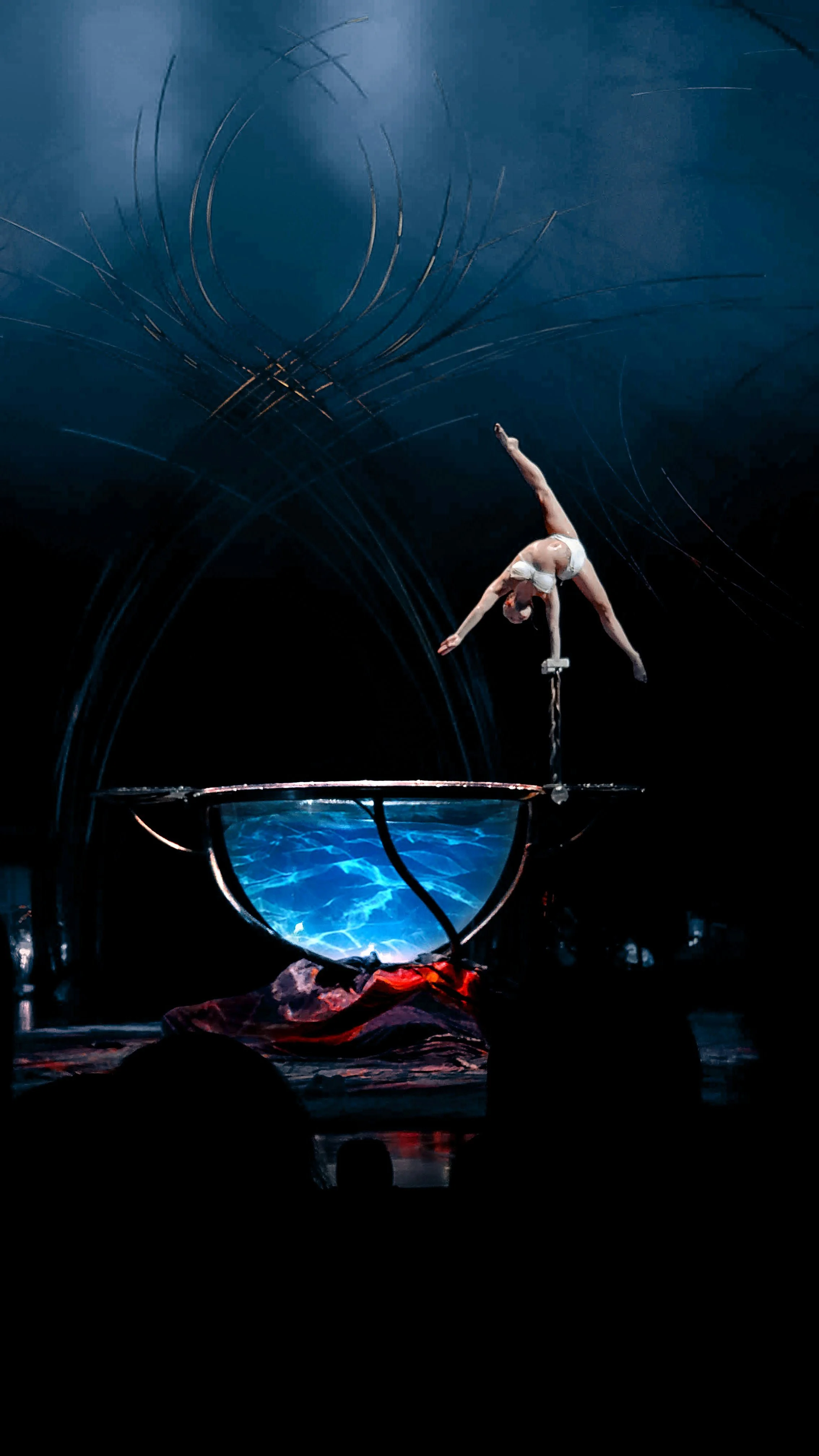 A female acrobat mid-performance balancing on a pole in a dark, illuminated stage with artistic light effects.