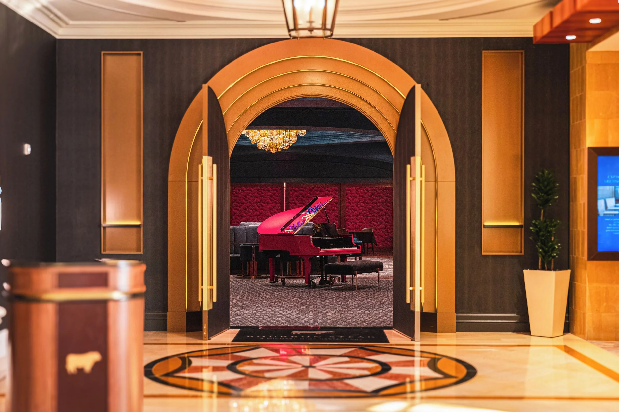 An elegant hotel lobby with a decorative tiled floor, a gold-trimmed arched doorway, and a pink grand piano in a lounge area with red tufted walls and chandeliers.